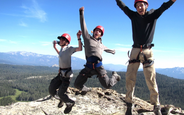 Three students wearing helmets and harnesses leap into the air in celebration. They appear to be on a summit, high above a wooded landscape. There are mountains in the background.